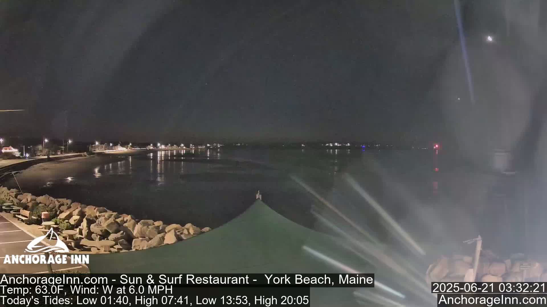 A nighttime view of a calm body of water with a shoreline featuring rocks and a partially visible awning, under a clear, dark sky.
