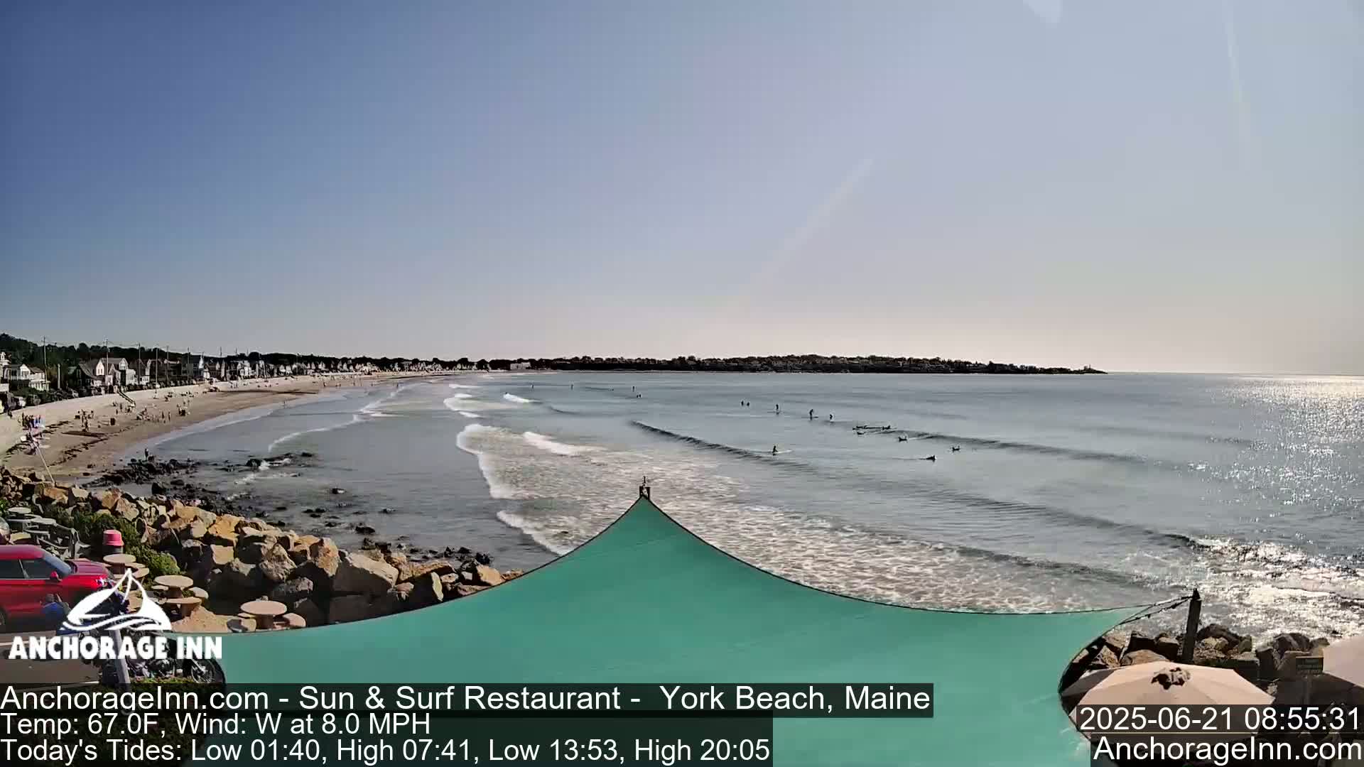 A sunny beach with small waves and several surfers, viewed from a rocky shoreline with a teal shade structure.