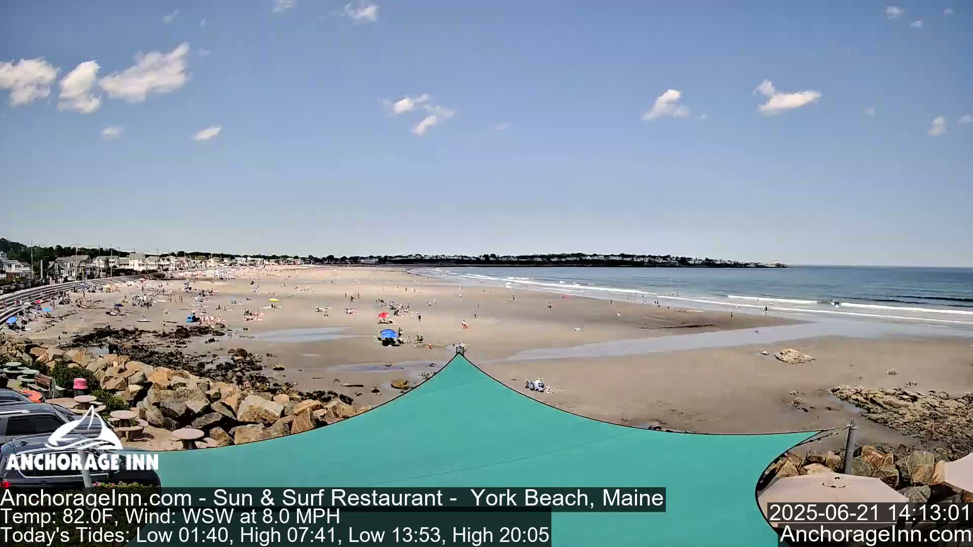 A sandy beach with many people, a calm ocean with small waves, and a partly cloudy sky is visible from a slightly elevated vantage point.