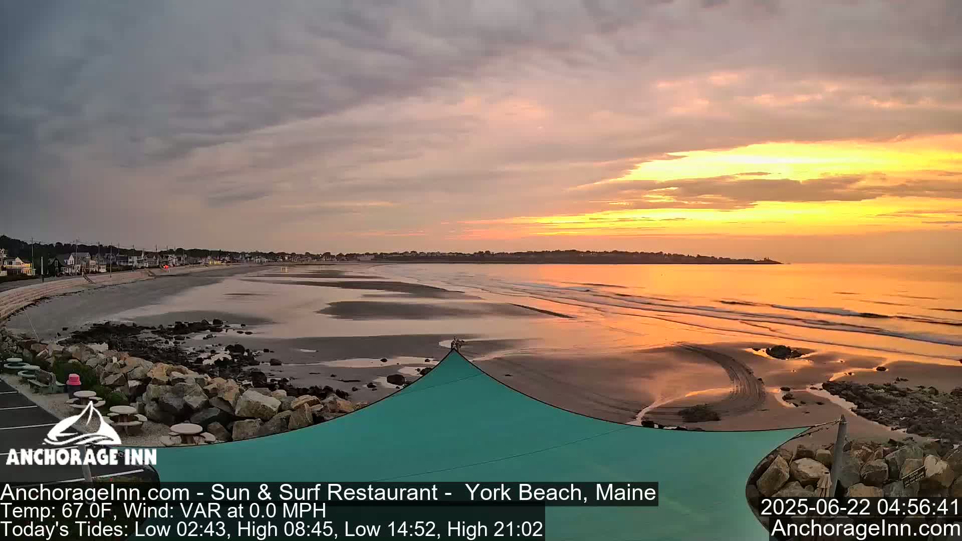 A wide sandy beach at sunrise, with calm ocean waves and a teal shade structure in the foreground.