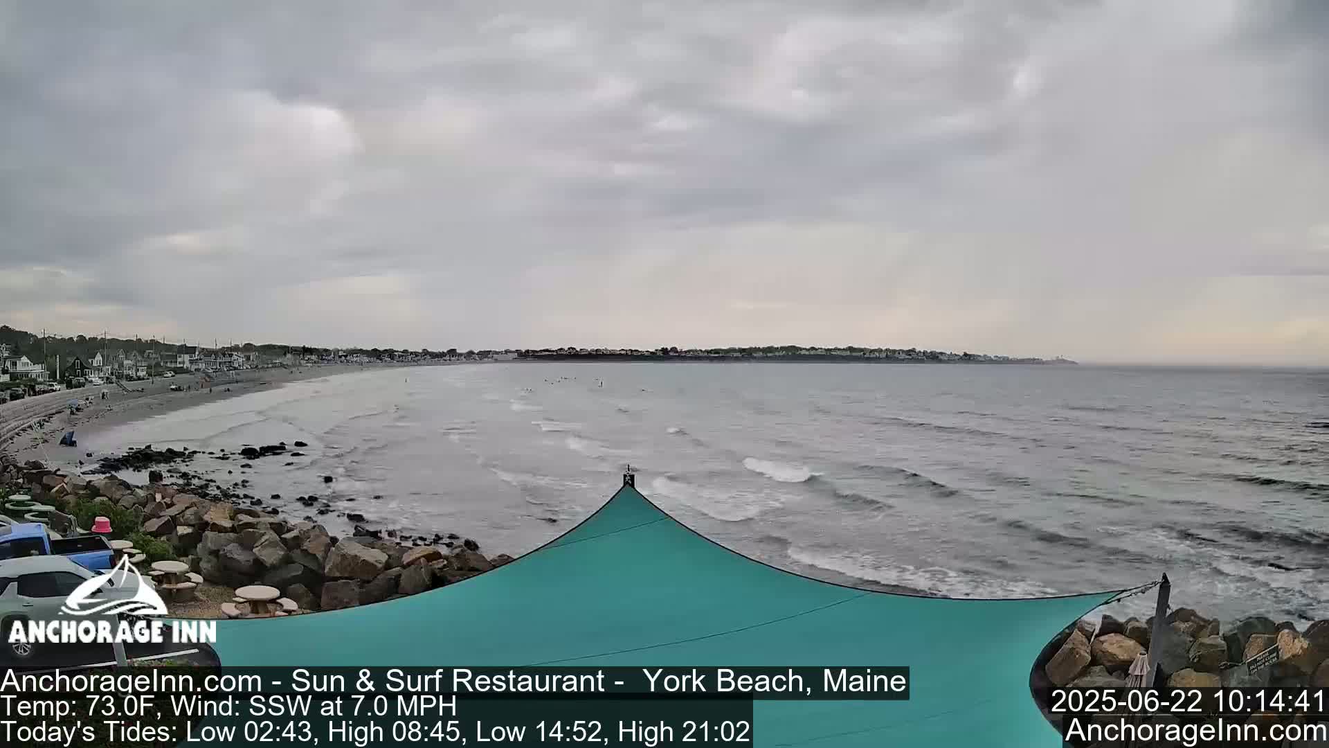 A mostly cloudy day shows a view of a beach with a calm ocean, small waves, and a teal shade structure partially visible in the foreground.