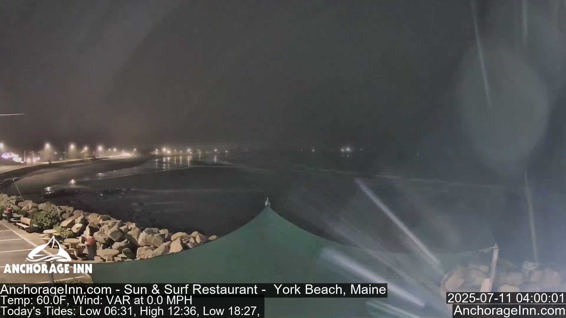 A nighttime view of a calm ocean with a rocky shoreline and a partially visible building under a hazy sky.