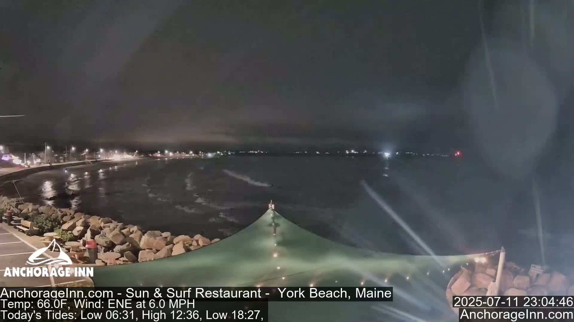 A nighttime view of a calm ocean with a rocky shoreline and a partially visible building under a hazy sky.