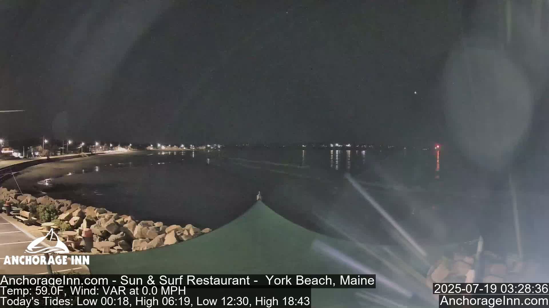 A nighttime view of a calm ocean reflecting lights from a town across the water, with a rocky shoreline in the foreground.