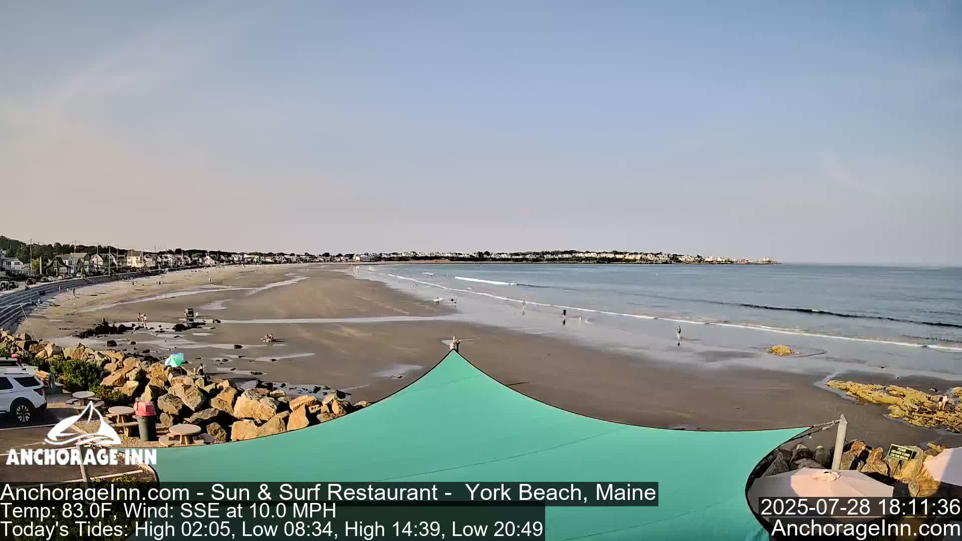 A sandy beach with a calm ocean and a few people is viewed from a rocky vantage point under a partly sunny sky.