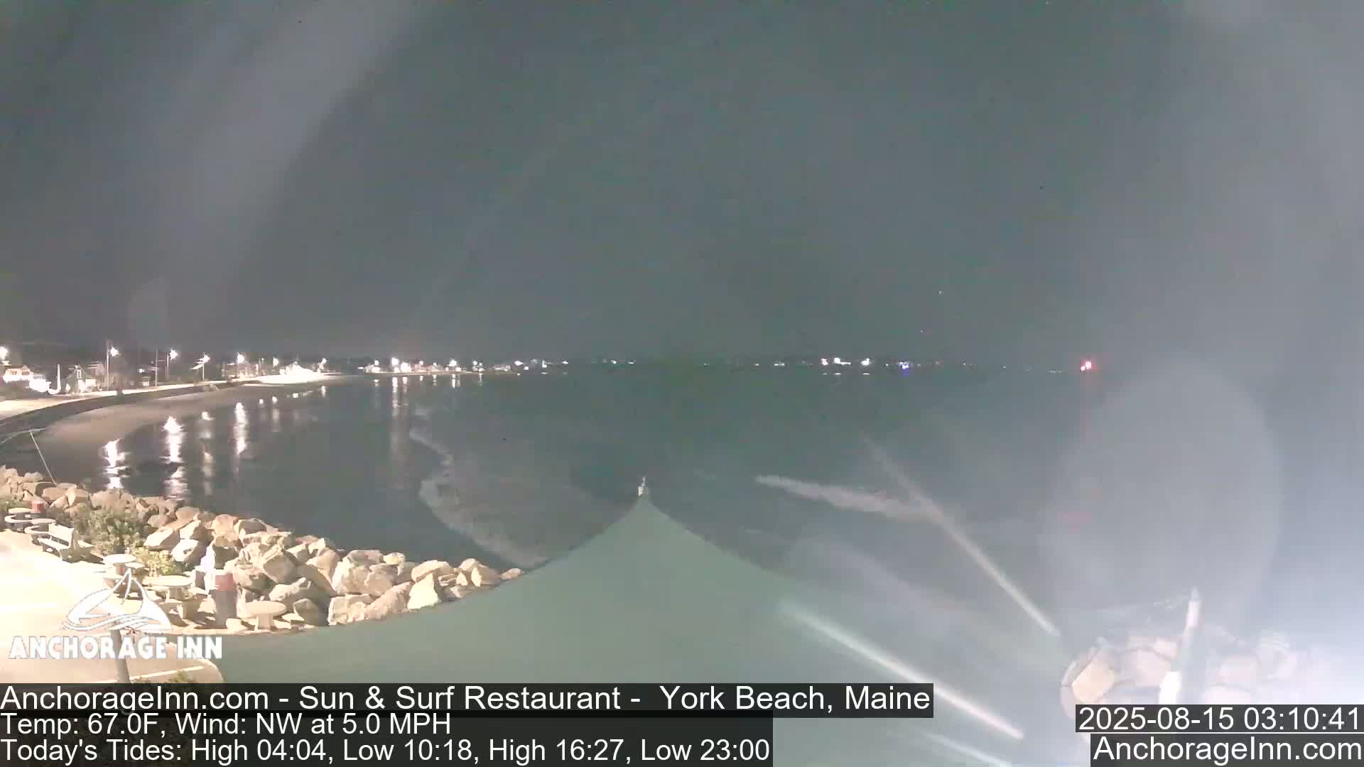 A nighttime view of a calm ocean next to a rocky shoreline, with lights visible in the distance under a dark sky.