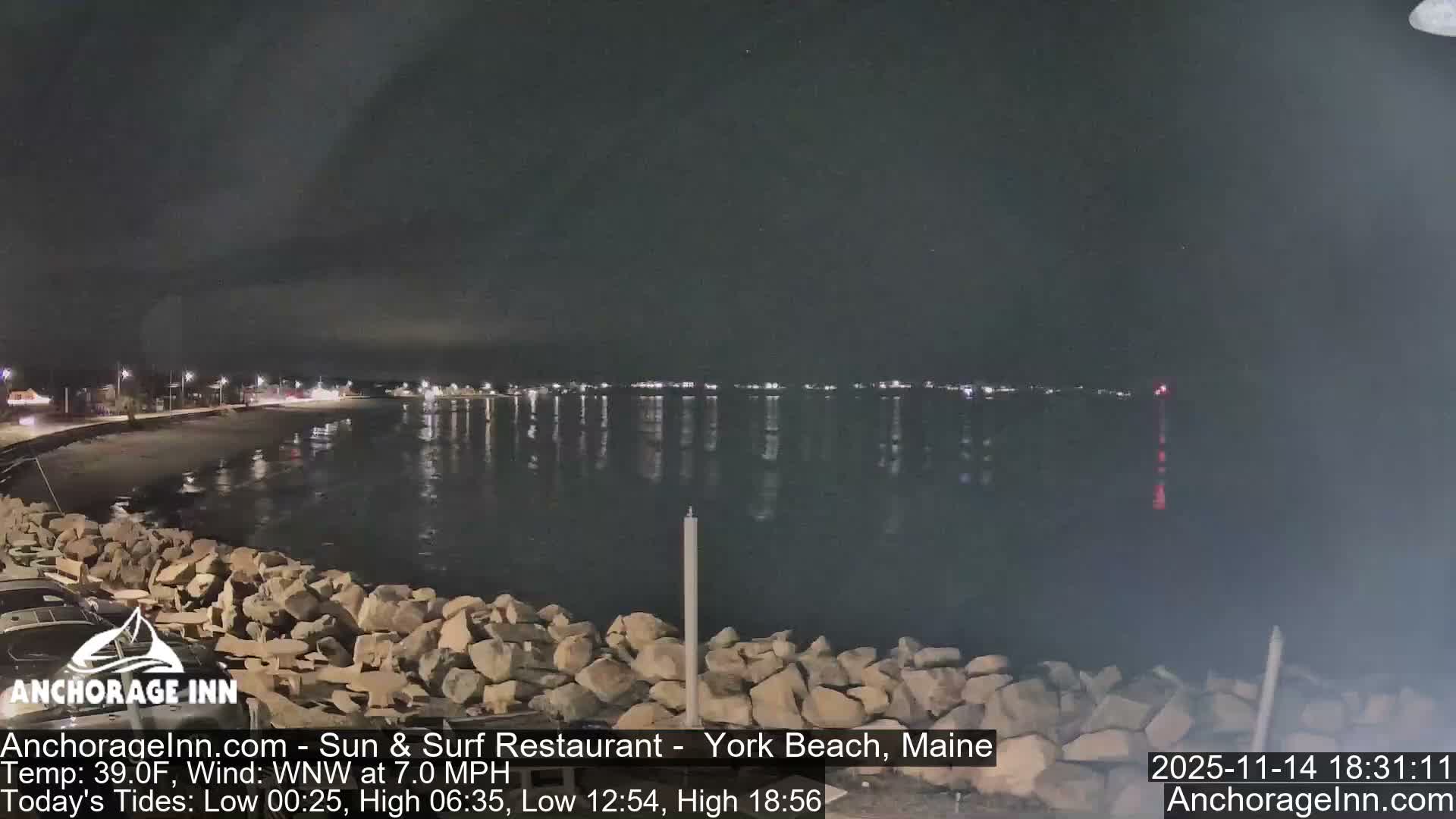 At night, a rocky shoreline borders a calm body of water reflecting distant lights from a town or opposing shore, all under a dark, cloudy sky.