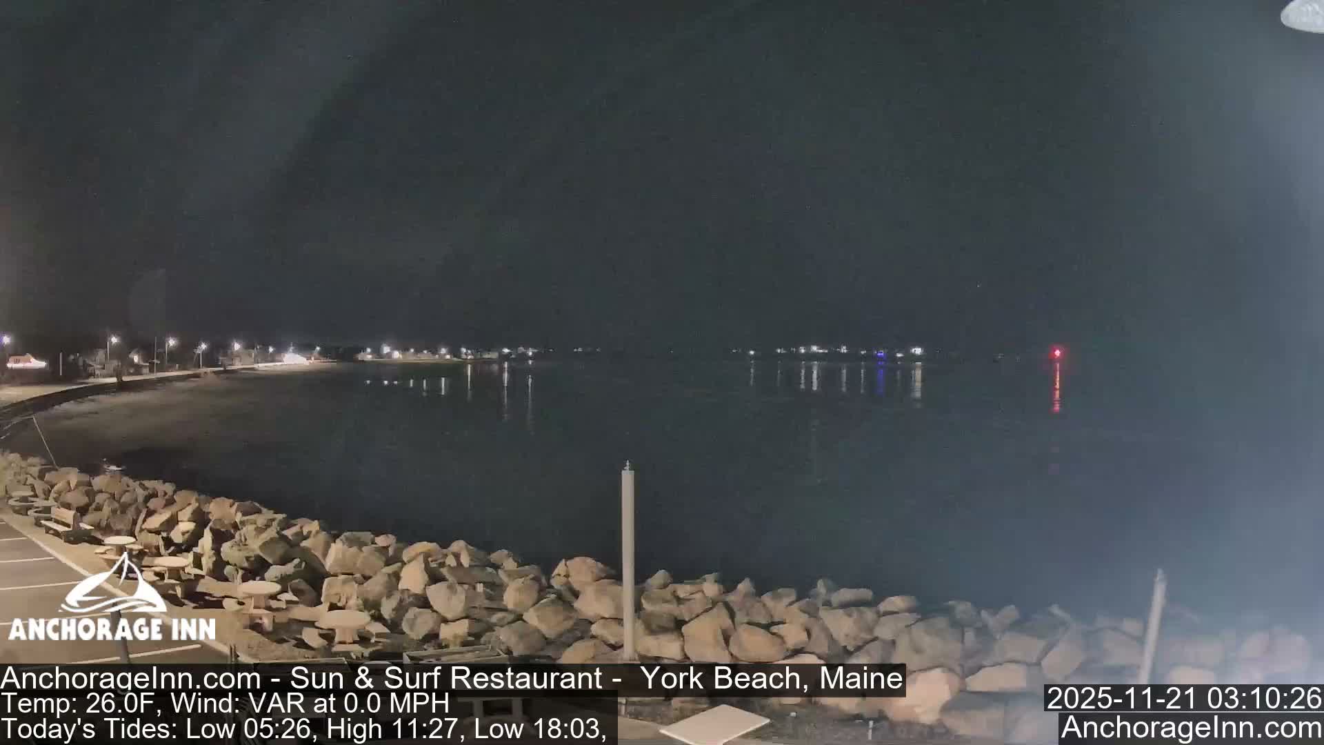 A dark night view reveals a rocky shoreline with a paved path and benches, illuminated by streetlights that reflect on the calm water of a bay, where distant town lights and a red buoy glow under a clear sky.