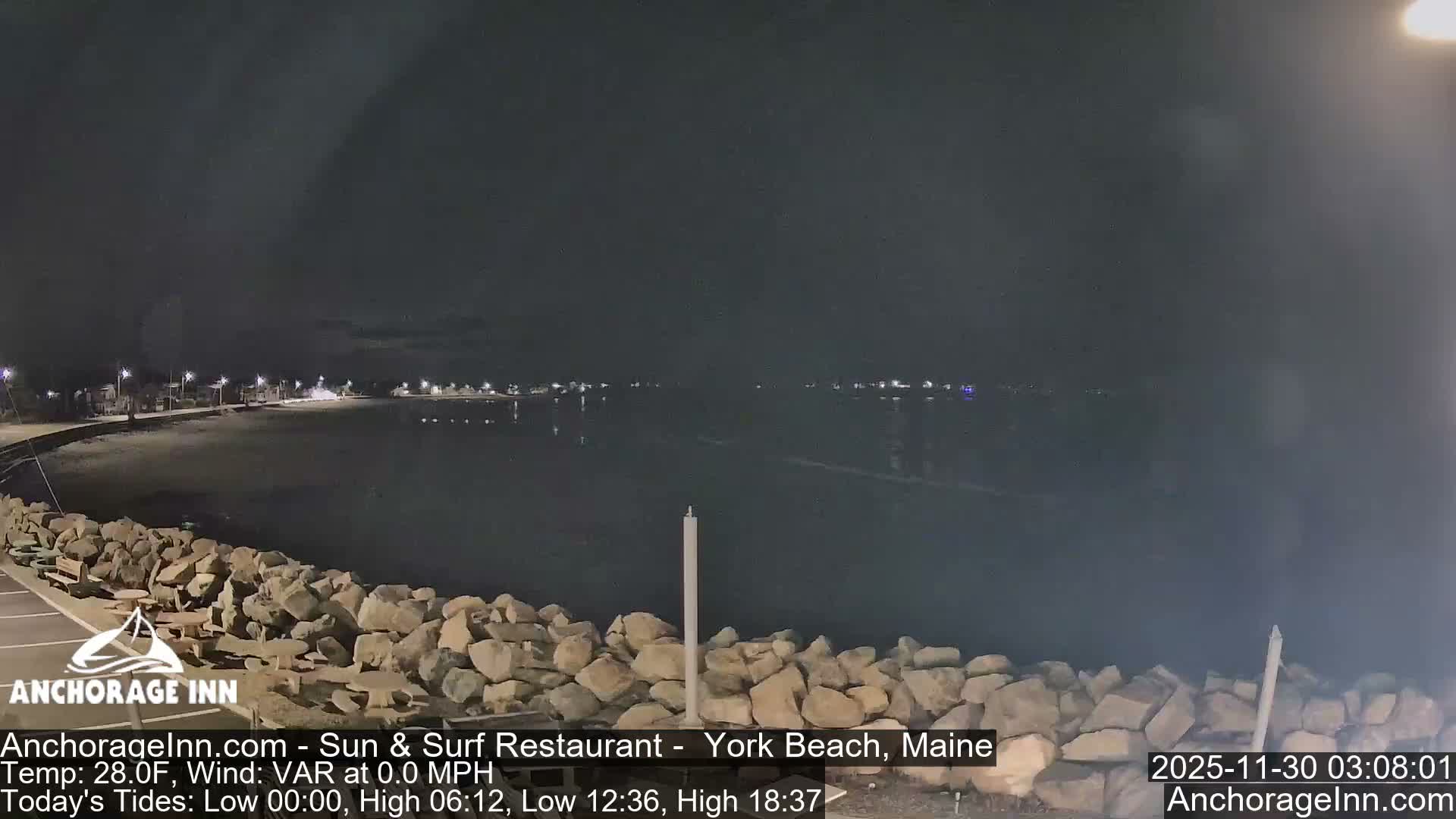 A dark, cloudy night view shows a coastline with a rocky barrier in the foreground, streetlights illuminating a curving road and buildings along a sandy beach to the left, and distant lights reflecting on the calm water to the right.