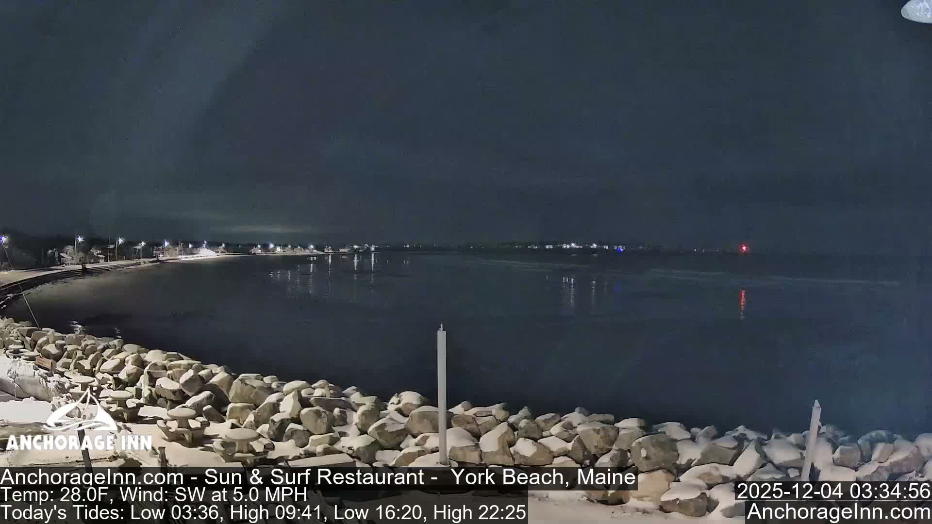 Under a dark, cloudy sky, a snow-covered rocky shoreline borders calm water reflecting distant coastal lights, with a solitary red beacon visible in the distance, indicating cold, wintry conditions.