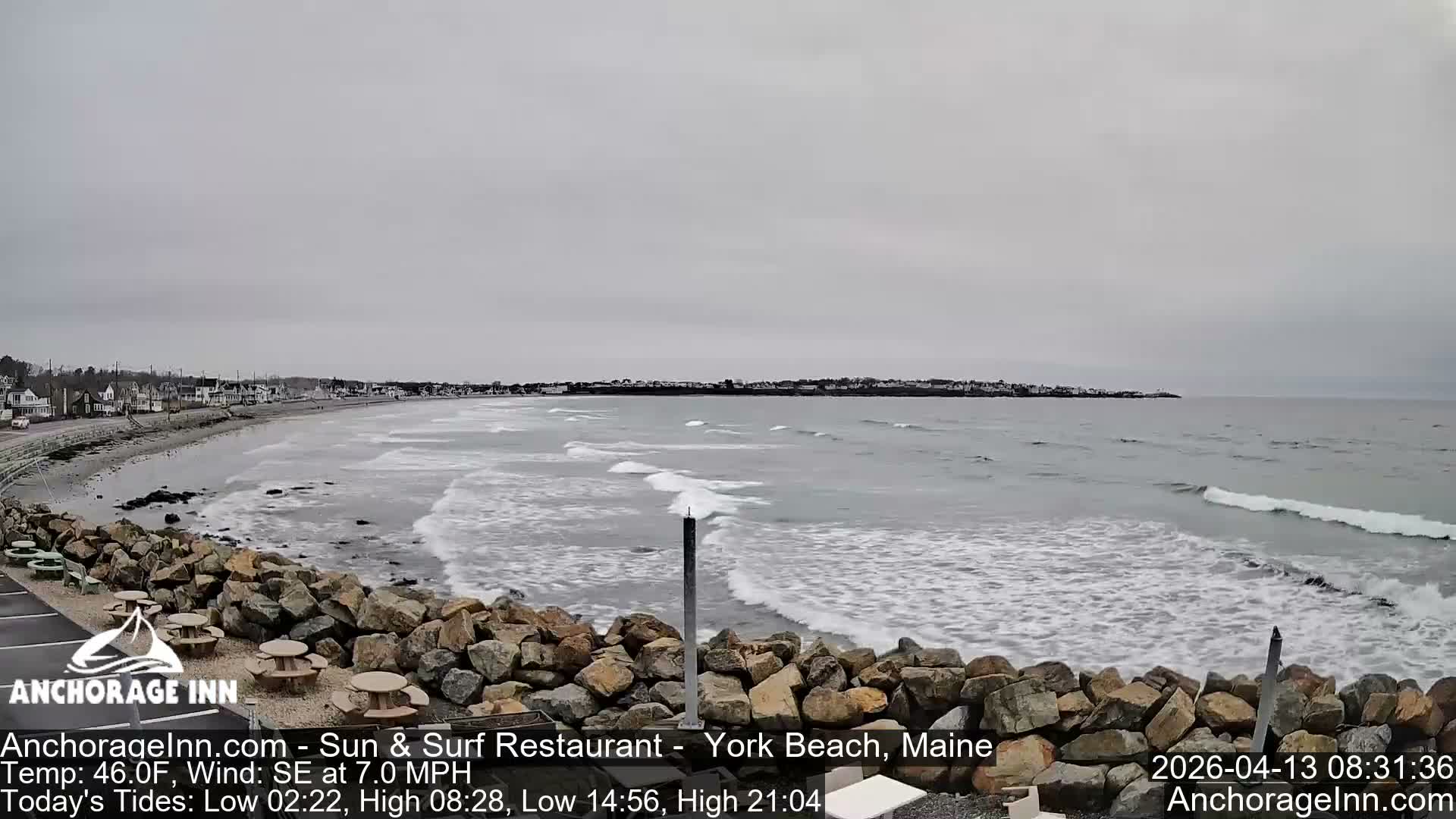 Under a dark, cloudy sky, a snow-covered rocky shoreline borders calm water reflecting distant coastal lights, with a solitary red beacon visible in the distance, indicating cold, wintry conditions.