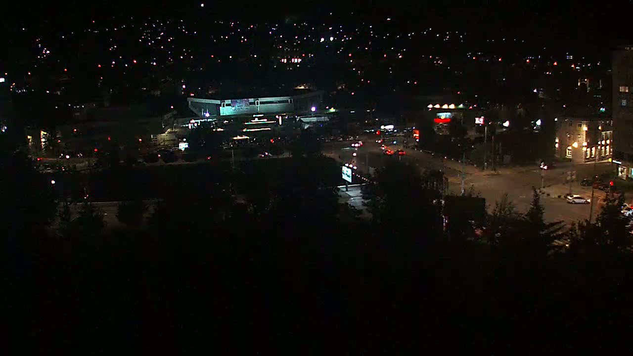 A nighttime, high-angle view of a city featuring a large building, roads with sparse traffic, and numerous small lights scattered across the landscape.