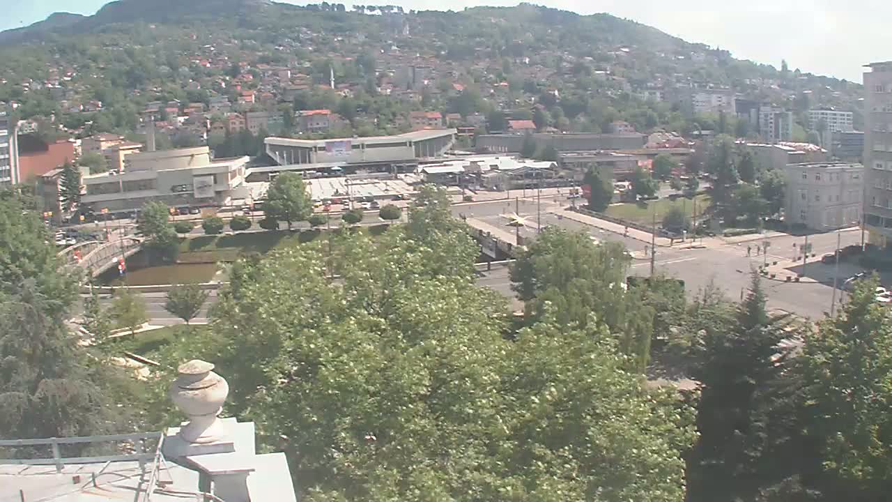 A high-angle view of a town on a sunny day, showing a river, bridge, buildings, and lush green trees.