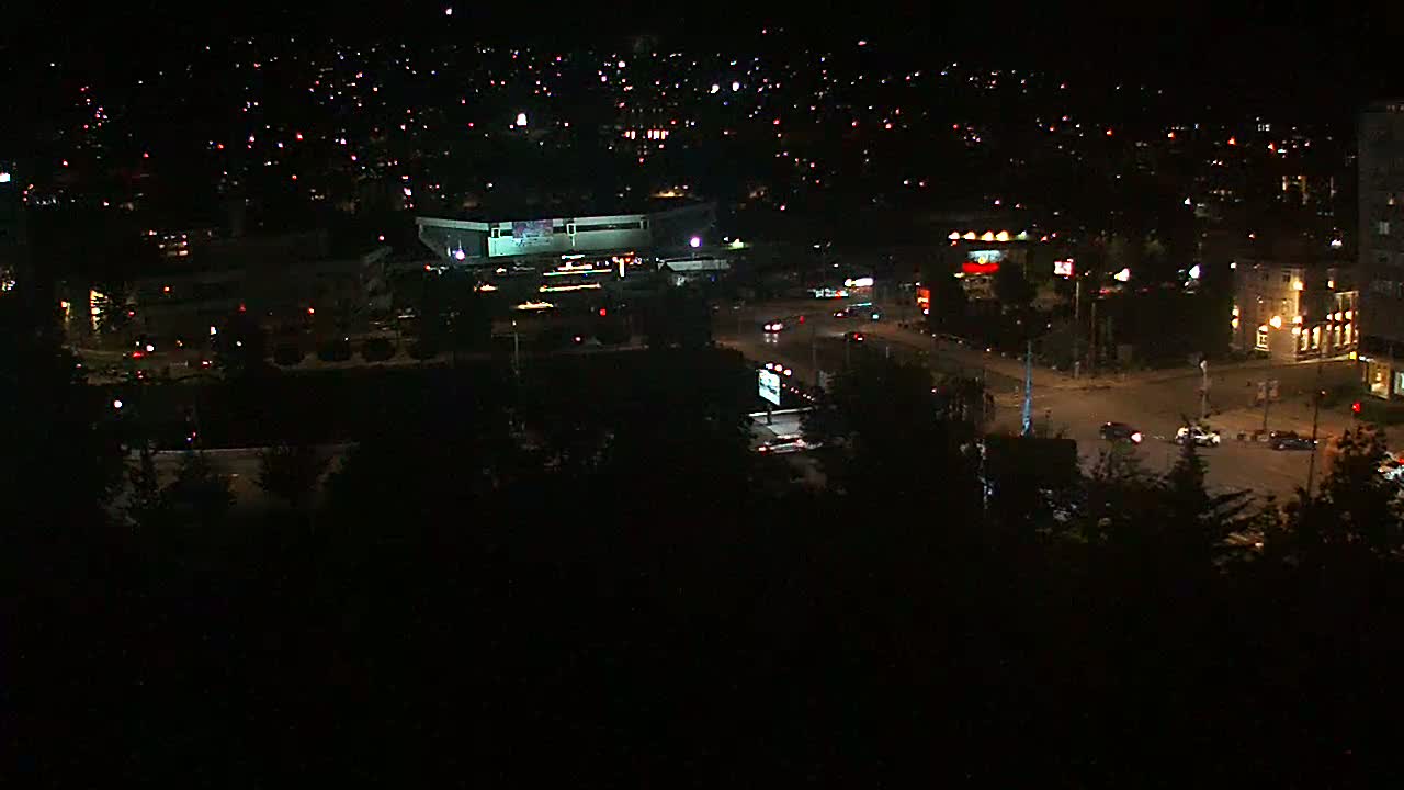 A nighttime, aerial view shows a city landscape with illuminated buildings, roads, and sparse vehicle traffic.