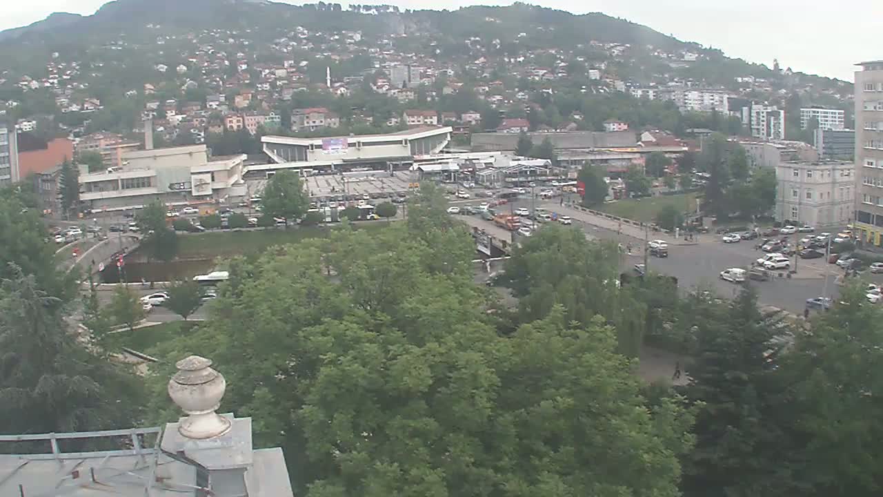 A high-angle view of a city on an overcast day shows a large building complex near a river, surrounded by green trees and a hillside of buildings in the distance.