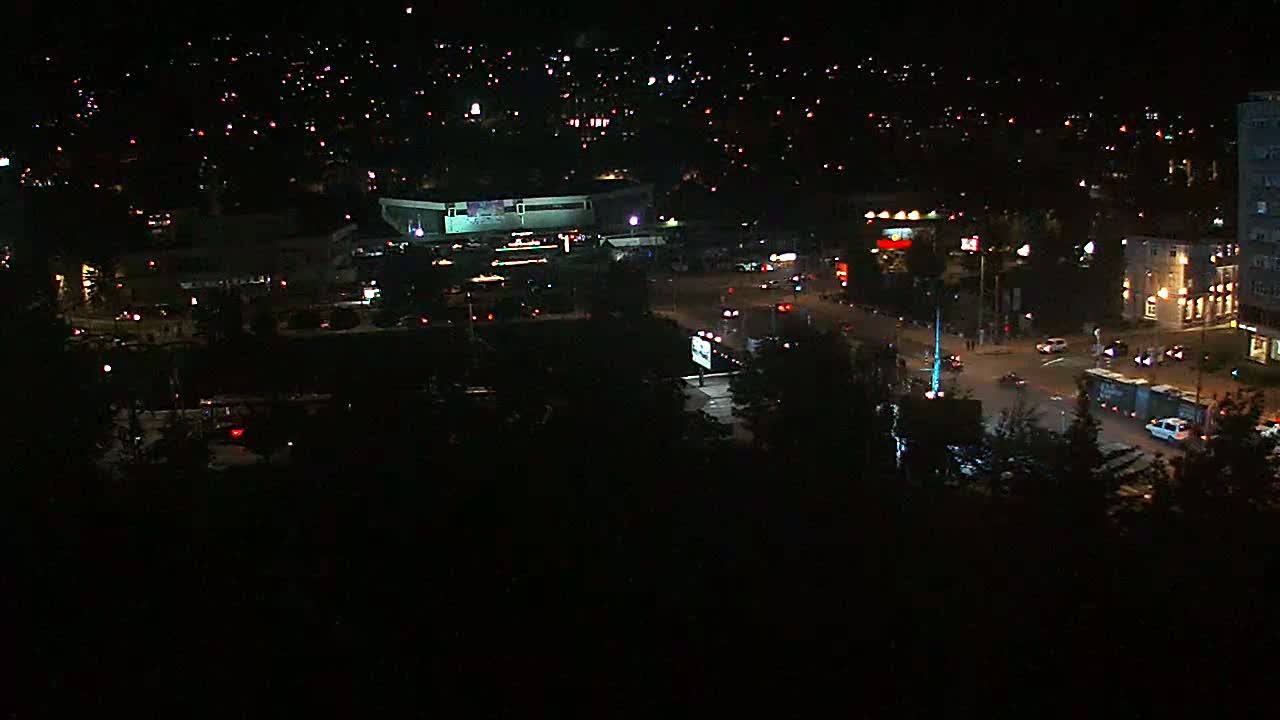 A nighttime aerial view shows a city intersection with sparse traffic, surrounded by dark buildings and trees under a clear sky.