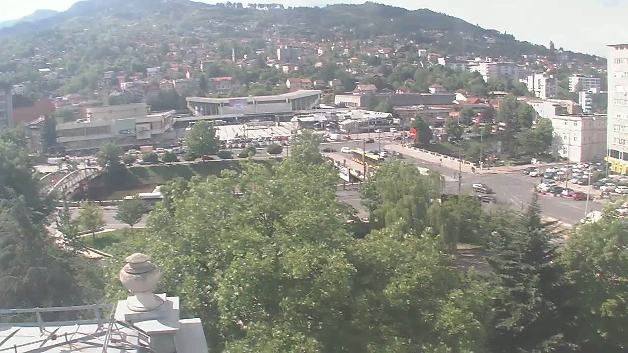 A high-angle view shows a city nestled in a valley under a clear sky, with lush green trees and a river flowing beneath a bridge.