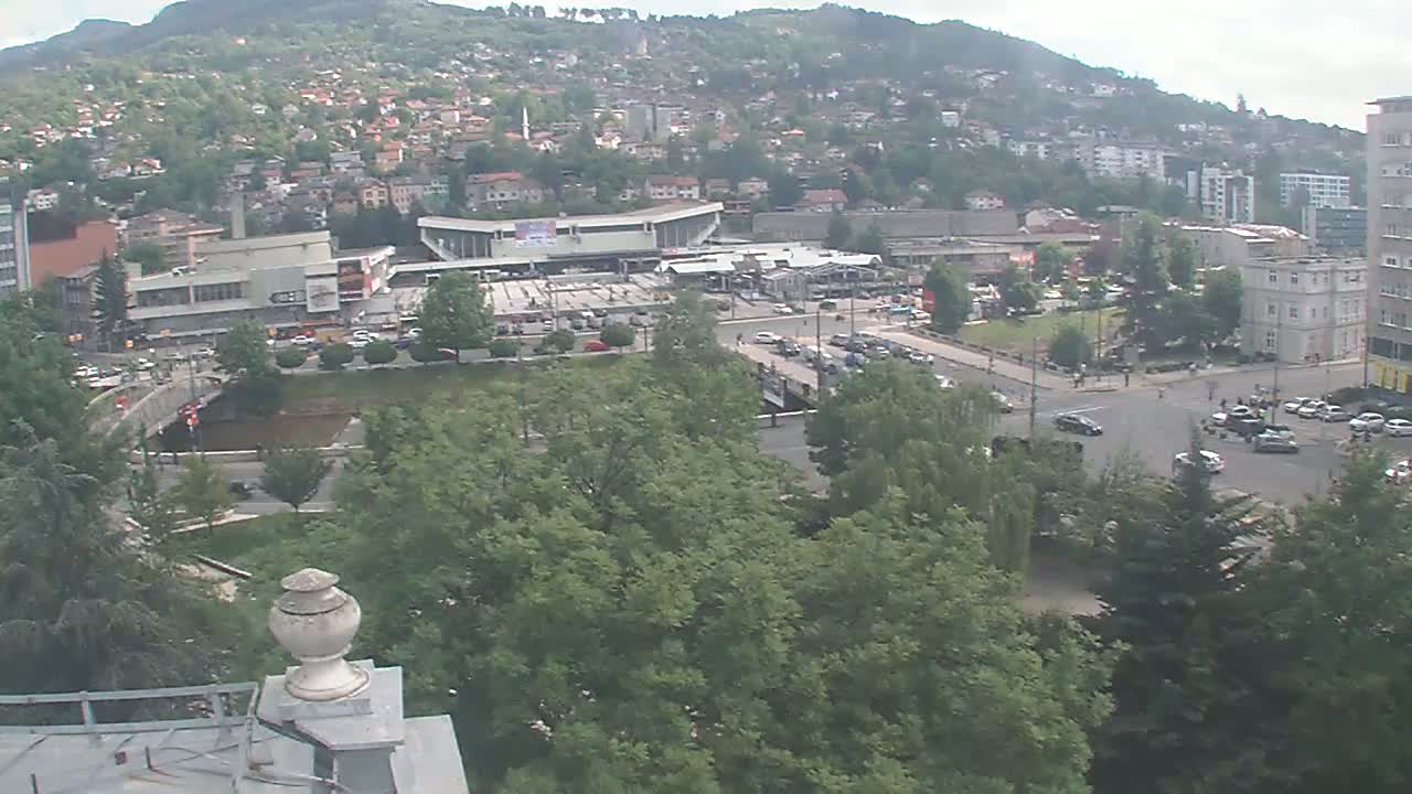 An overcast day reveals a city scene with a river, bridge, buildings, and lush green trees, nestled against a hillside.