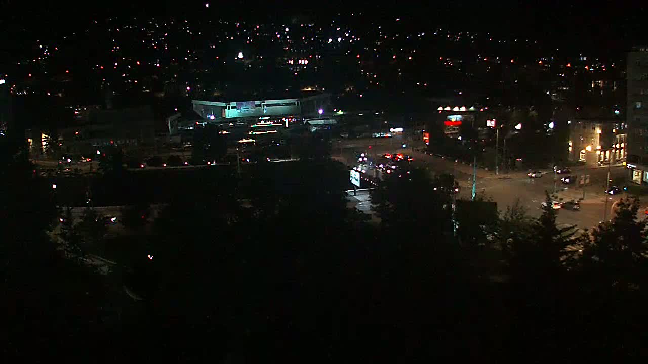 A nighttime aerial view shows a city intersection with sparse traffic, a large building, and surrounding buildings with scattered lights.