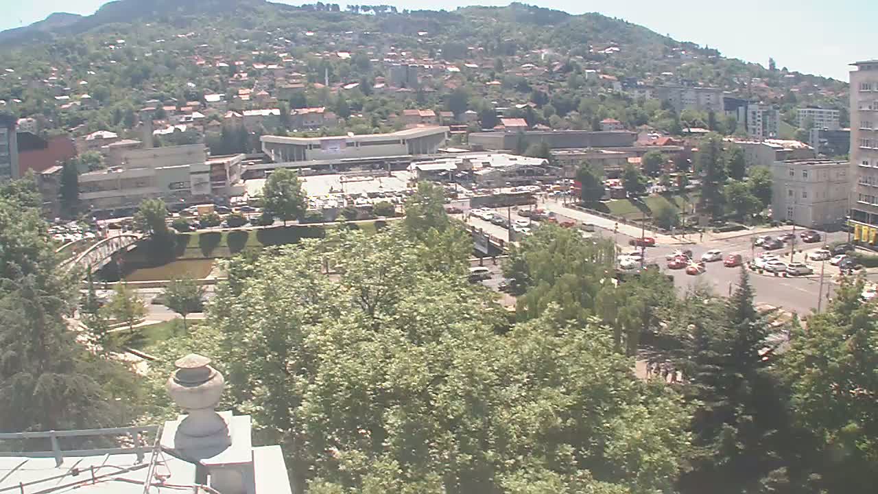A high-angle, long shot on a sunny day shows a city nestled in a valley, with a river, bridge, and buildings visible amidst lush green trees.