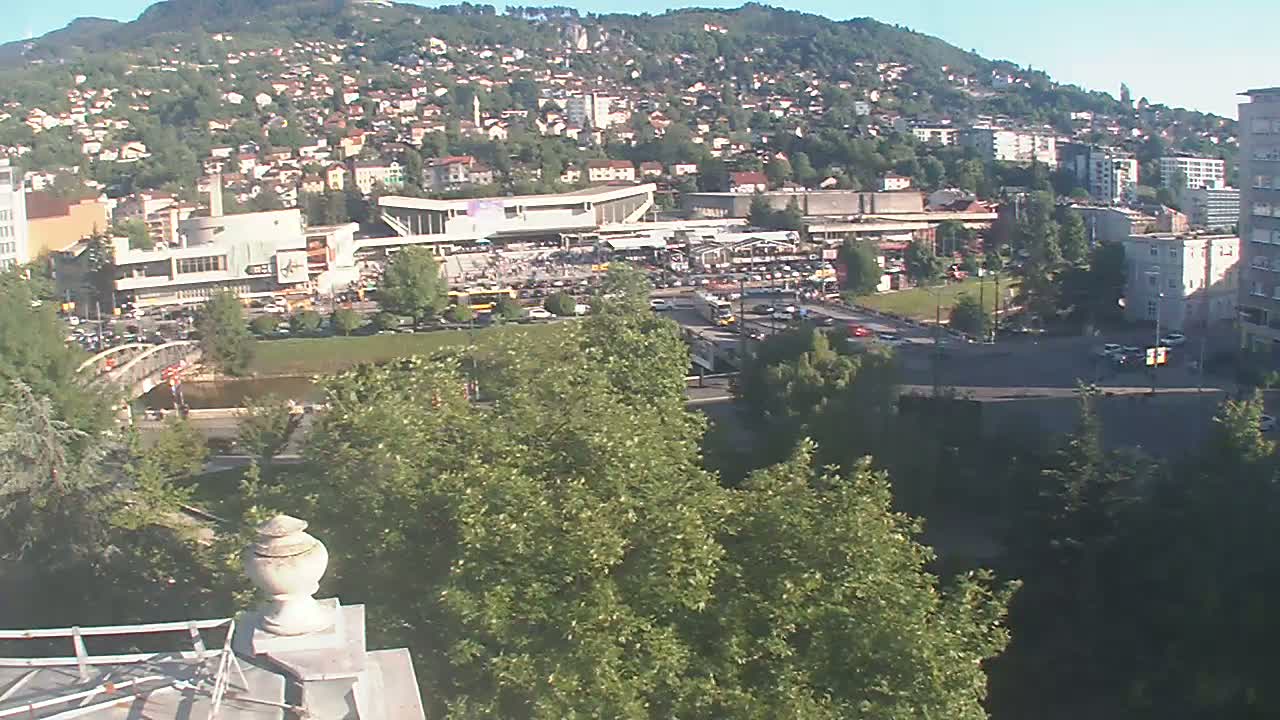 A high-angle view on a sunny day shows a city nestled against a hillside, with a large plaza and bus station in the foreground, and a river flowing nearby.