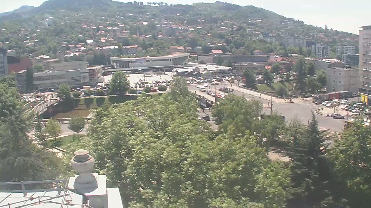 A high-angle, long shot on a sunny day shows a city nestled in a valley with green hills in the background, featuring buildings, roads, trees, a river, and a bridge.