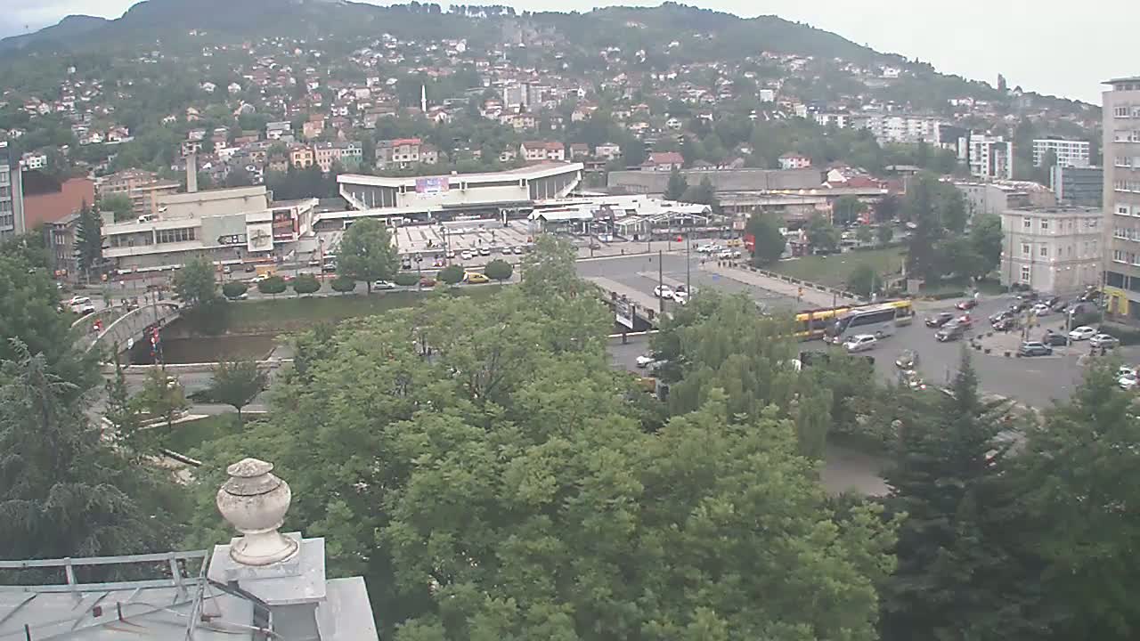 An overcast day reveals a city scene with a bus station, bridge, and river, surrounded by lush green trees and buildings on a hillside.