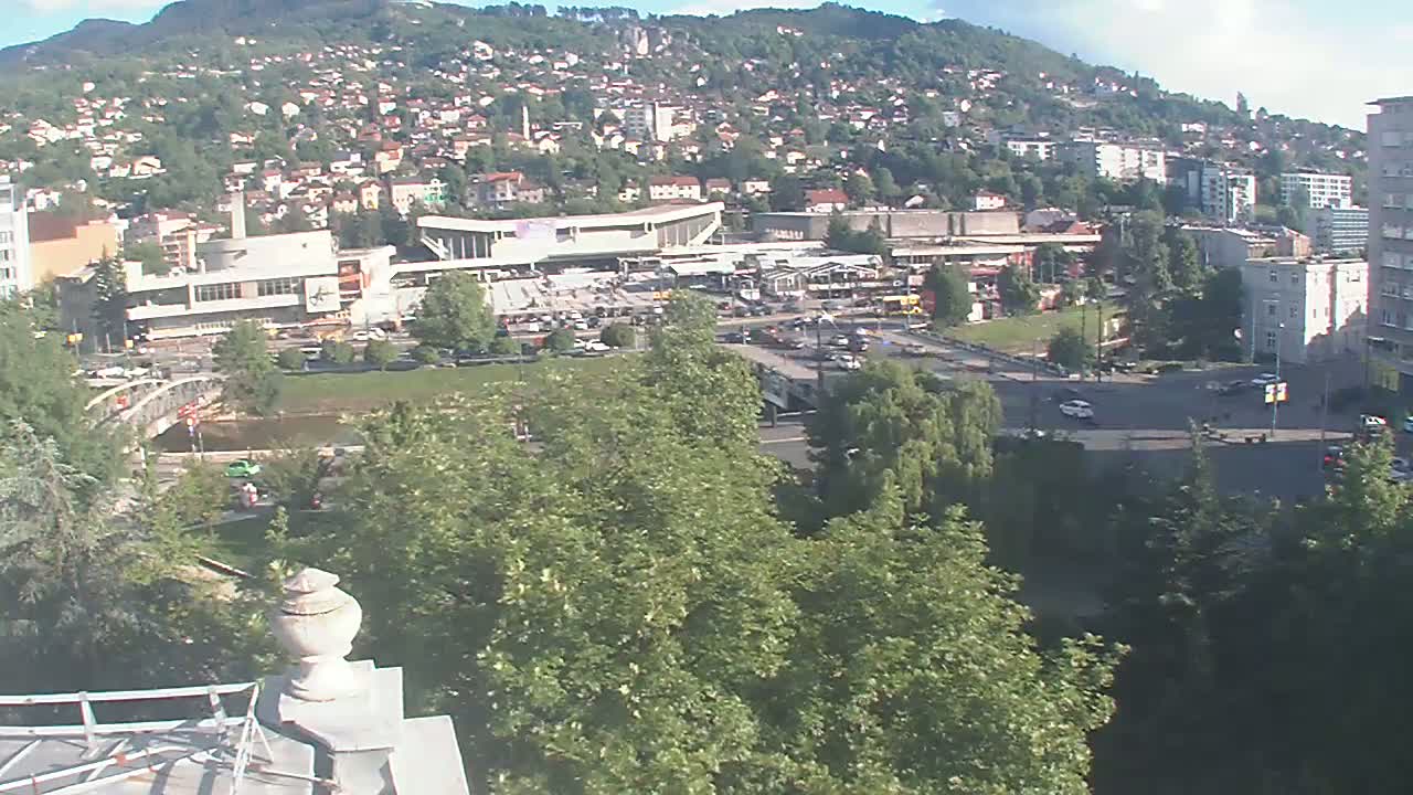 A high-angle view of a city nestled against a hillside on a partly sunny day, showing a river, bridge, buildings, and lush green trees.