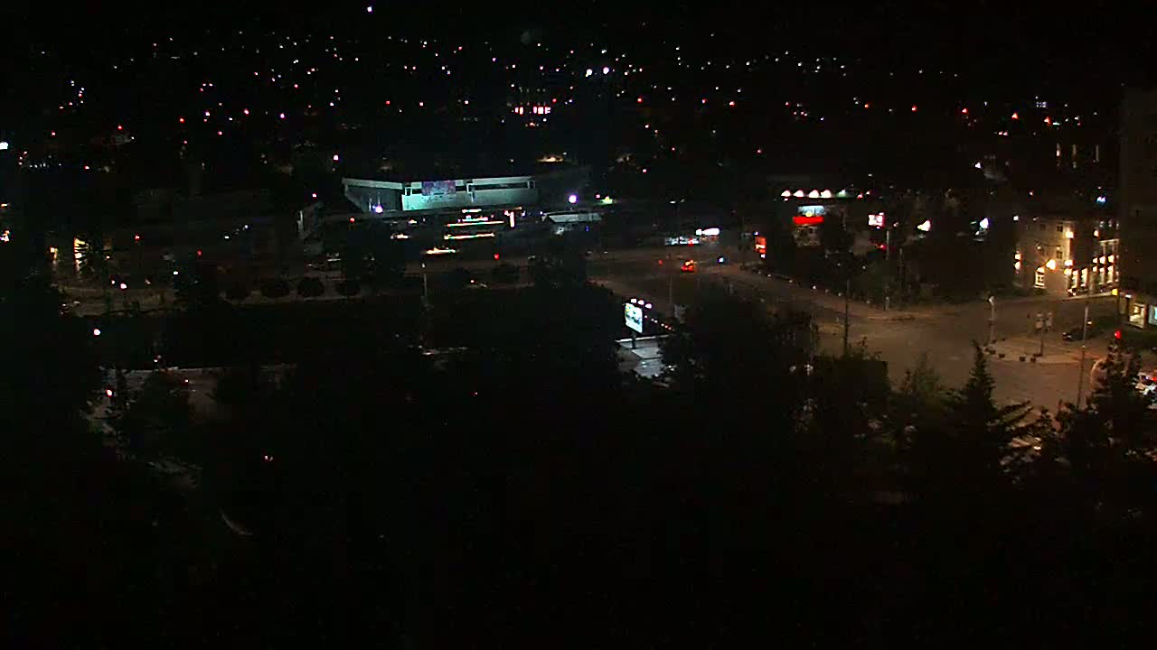 A nighttime aerial view shows a city intersection with sparse traffic, buildings with illuminated windows, and a large dark area, possibly a park, with surrounding streetlights.