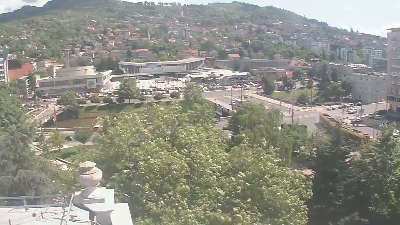 A sprawling city scene on a sunny day, featuring a large building complex near a river, surrounded by lush green trees and hills dotted with houses in the background.