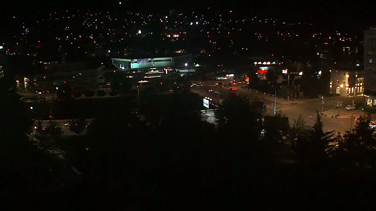 A nighttime aerial view of a city shows a large building, roads with cars, and numerous streetlights illuminating the surrounding area.