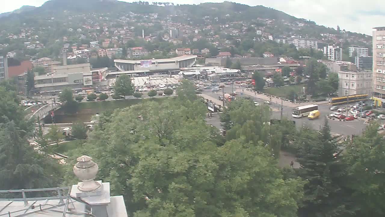 An overcast day reveals a city scene with a river, bridge, buildings, vehicles, and lush green trees, nestled against a hillside.
