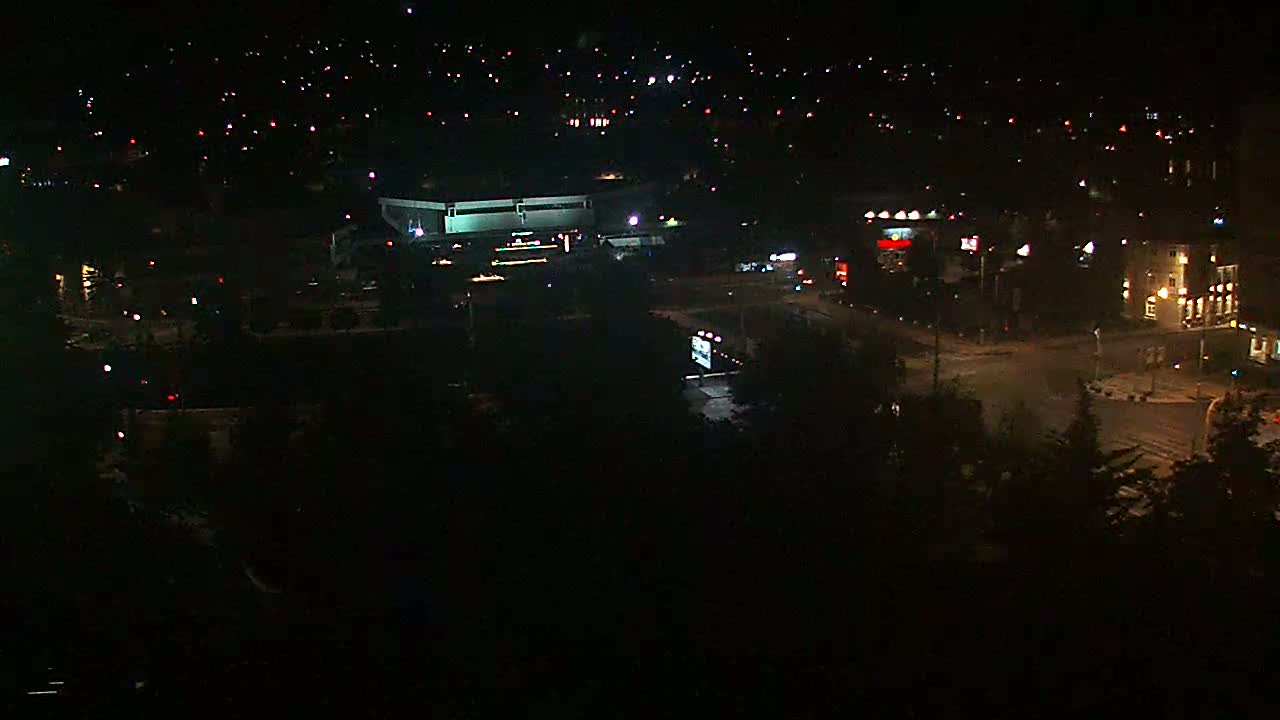 A nighttime aerial view shows a city intersection with buildings and sparse vehicle traffic under a clear sky.