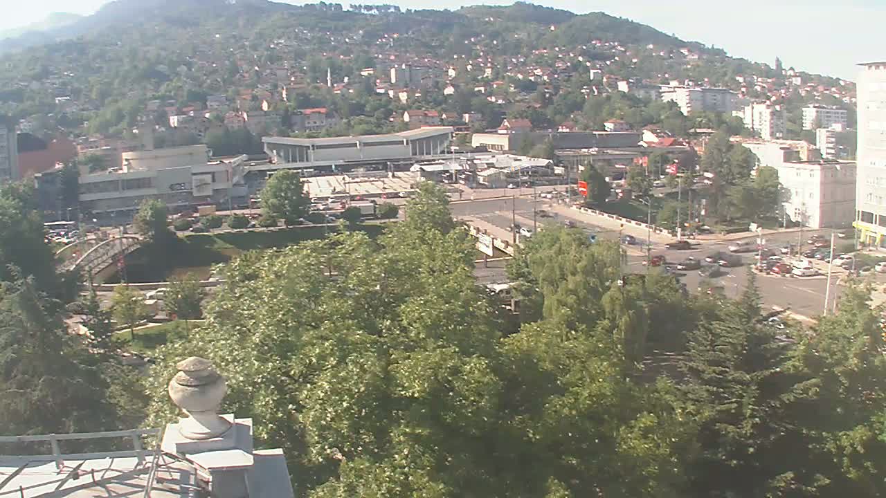 A high-angle view on a sunny day shows a city nestled in a hillside, with a large building and a bridge visible, surrounded by lush green trees.