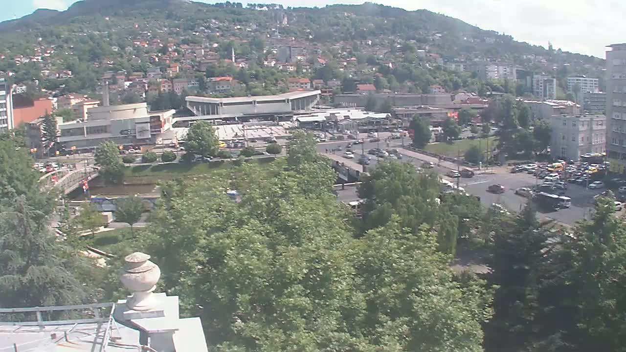 A high-angle, long shot on a sunny day shows a city nestled against a hillside, with a river, bridge, and numerous buildings, including a large, flat-roofed structure that appears to be a bus station.