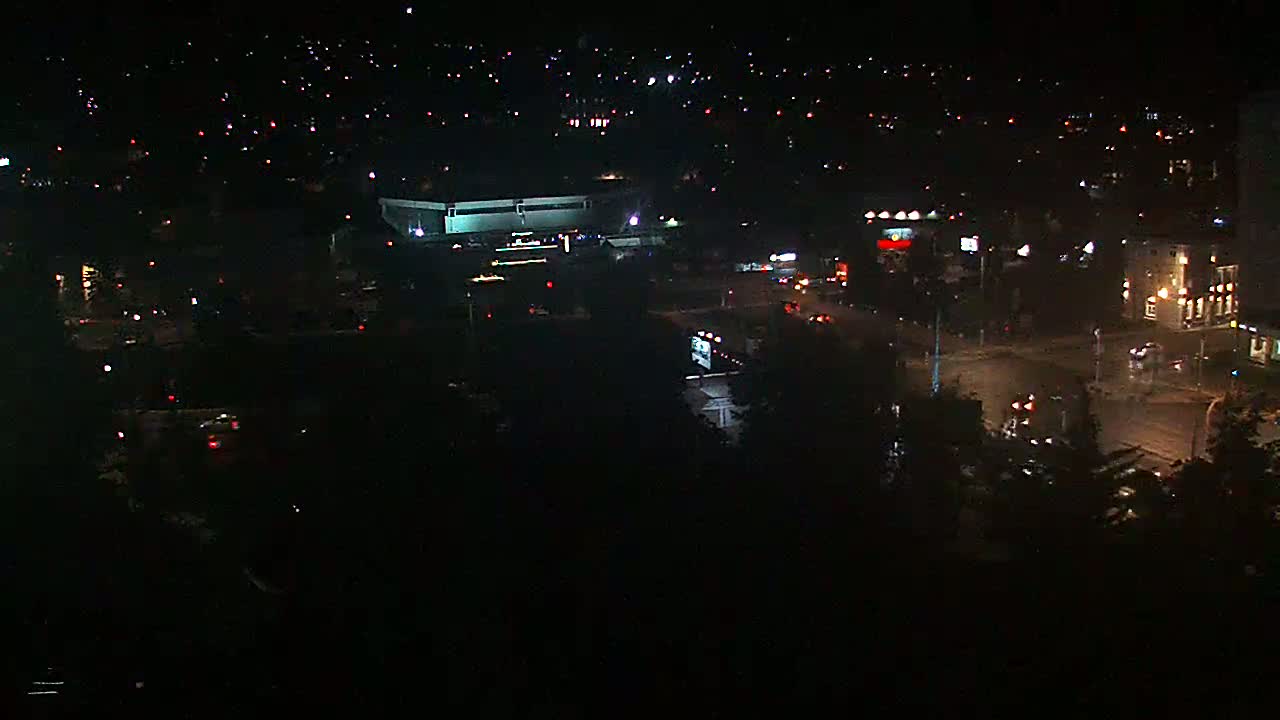 A nighttime aerial view of a city intersection with sparse traffic, showing illuminated buildings and a scattering of house lights in the distance.