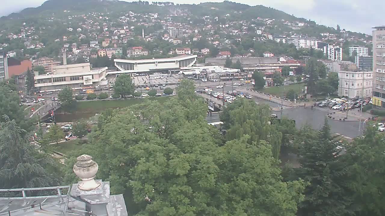 An elevated view of a city nestled against a hill on an overcast day, showing a large building complex, a bridge over a river, and surrounding greenery.