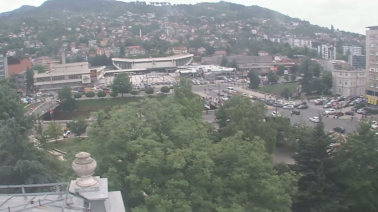 An overcast day reveals a city scene with a large building complex near a bridge and river, surrounded by lush green trees and numerous parked and moving cars.