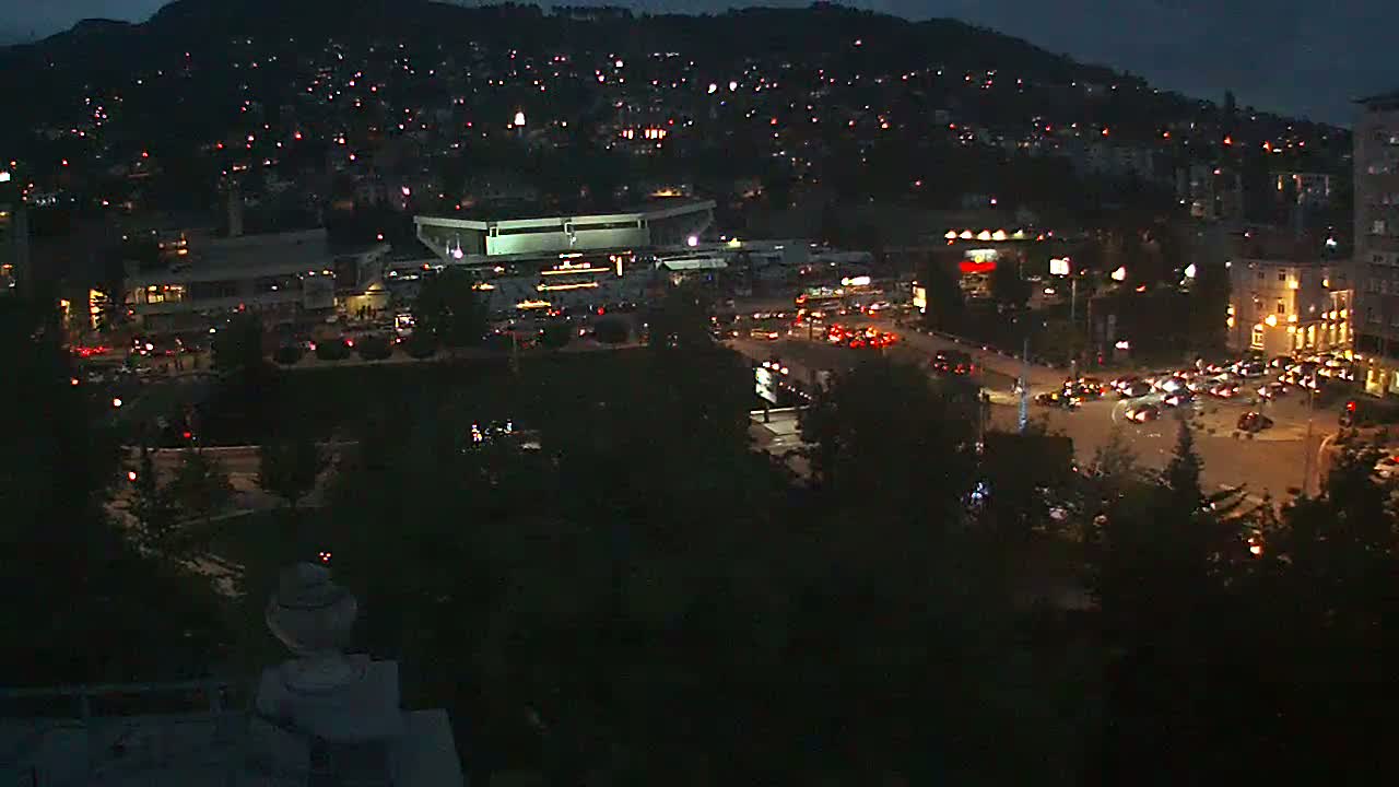 A nighttime aerial view shows a city with illuminated buildings, streets filled with cars, a large stadium, and a hill dotted with houses in the background under a dark sky.