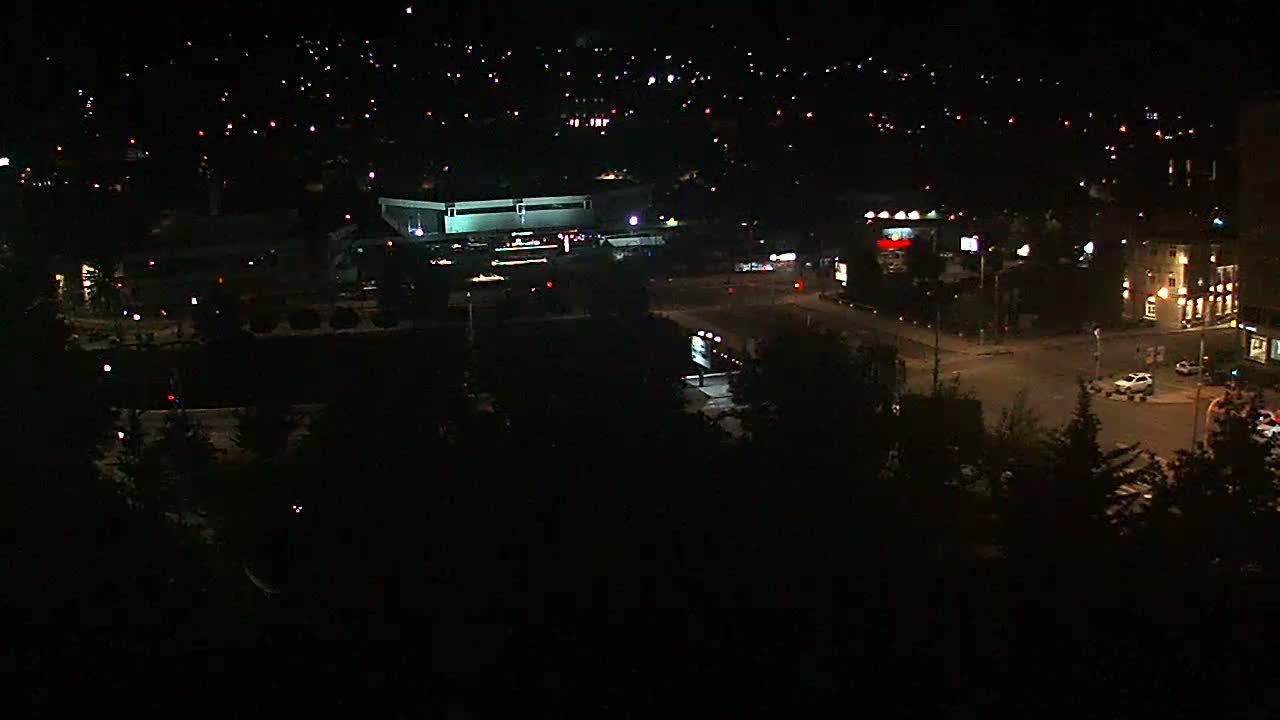 A nighttime aerial view of a city intersection with buildings and sparse traffic, illuminated by streetlights and house lights.