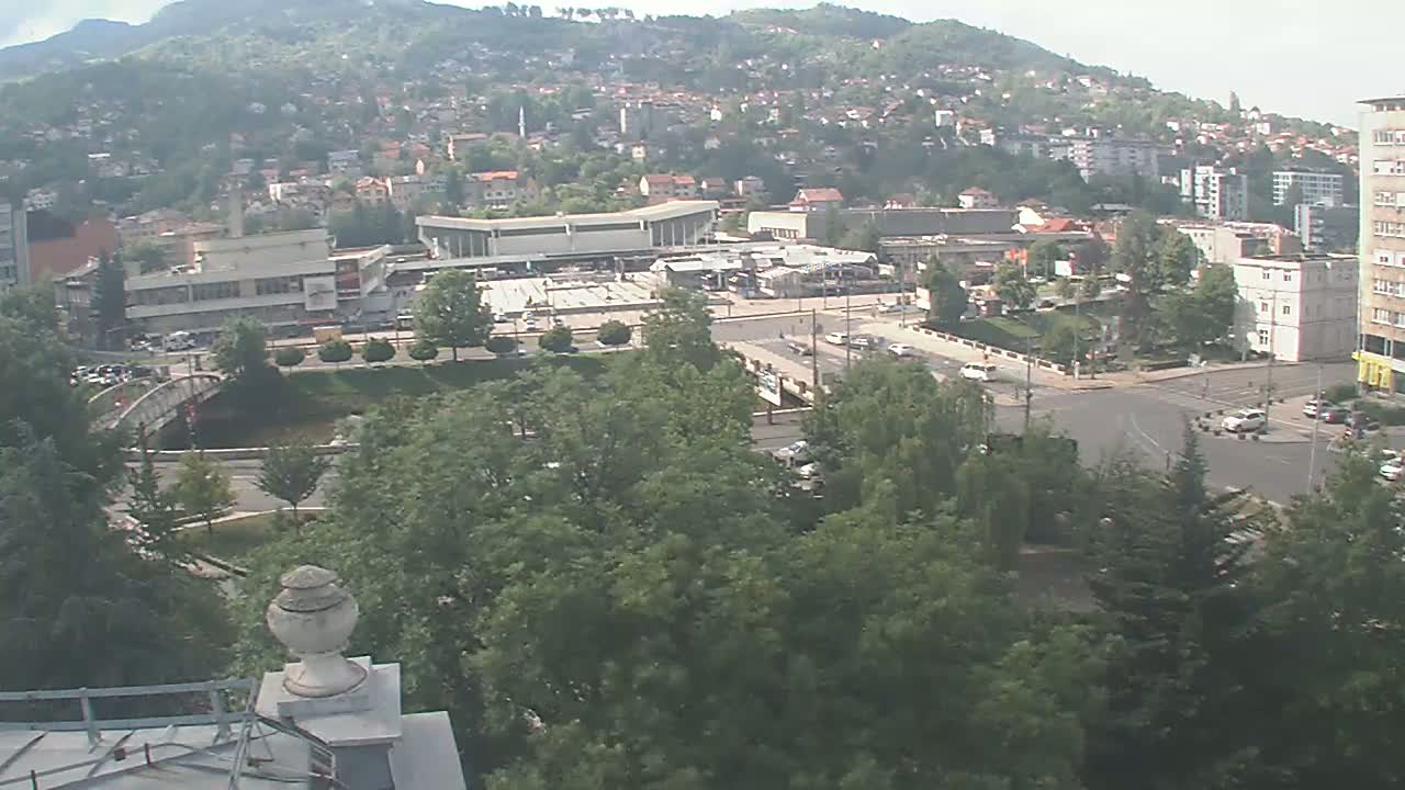 An elevated view of a city on a hazy day shows a large plaza with a bus station, a bridge over a waterway, and surrounding buildings nestled against a hillside.