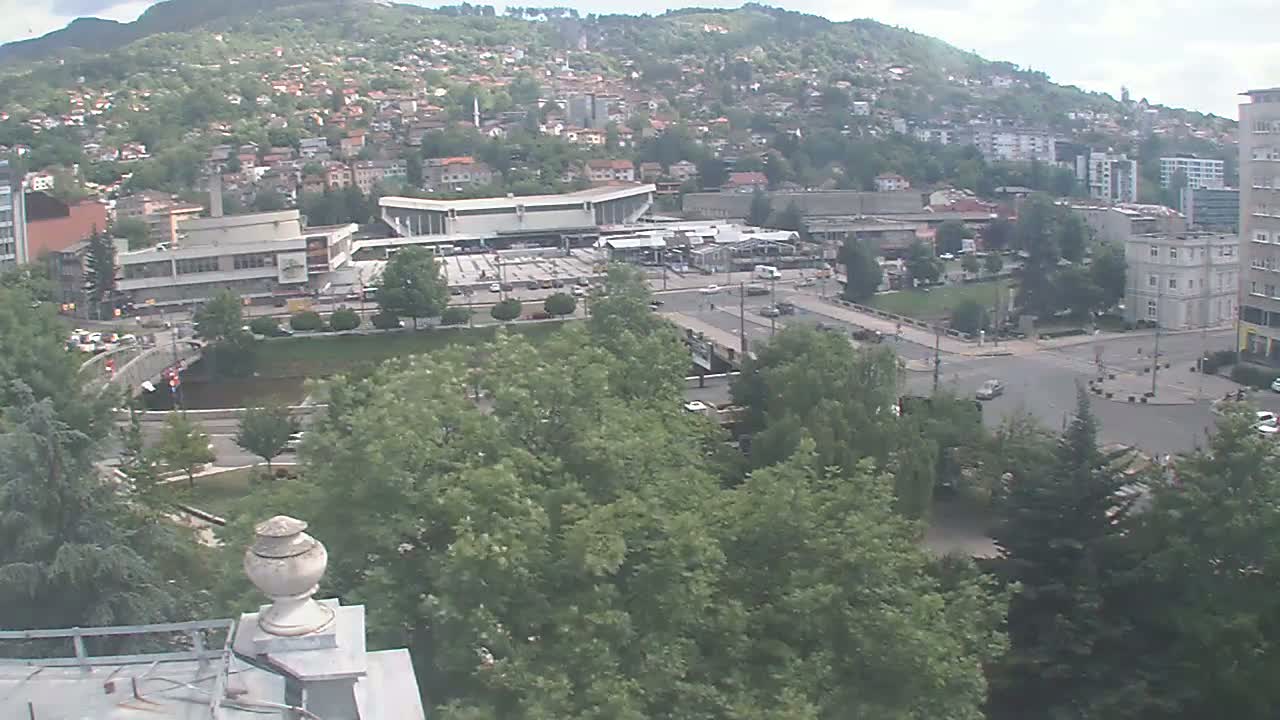 A high-angle view shows a city square with a bus station, buildings, trees, and a bridge on a partly cloudy day.