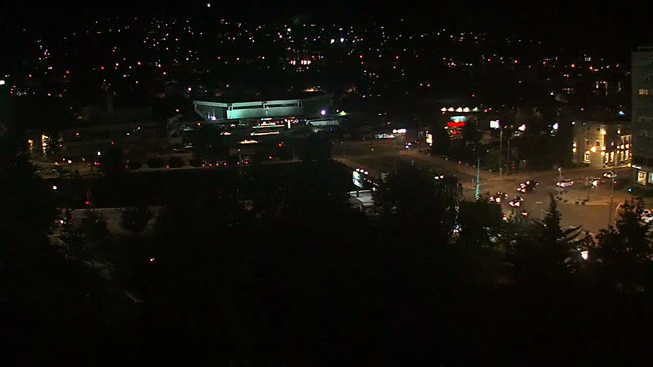 A nighttime aerial view shows a city intersection with several vehicles, buildings, and streetlights illuminated against a dark sky.