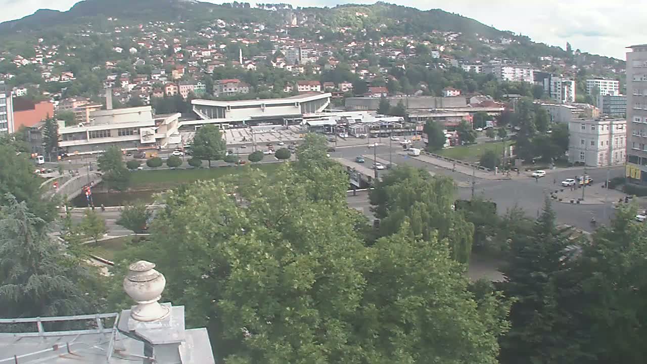 A high-angle view of a city on a partly cloudy day, showing a bus station surrounded by buildings and green spaces, with a hill in the background.