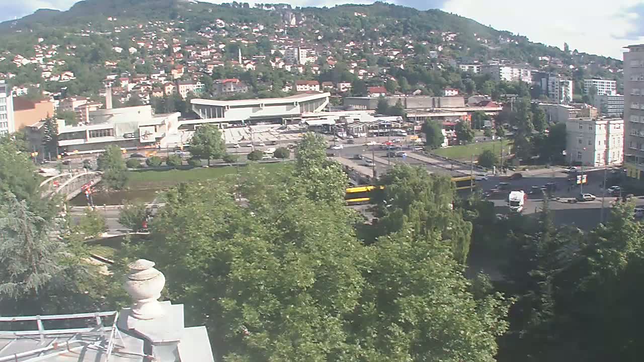 A high-angle view on a partly cloudy day shows a city scene with a large building, a bridge, a river, numerous vehicles, and lush green trees, all nestled against a hillside covered in houses and vegetation.