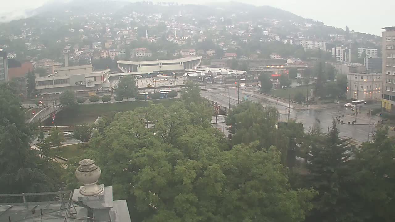 A rain-soaked city scene shows a sprawling urban area with buildings, roads, a river, and lush green trees, viewed from an elevated perspective.