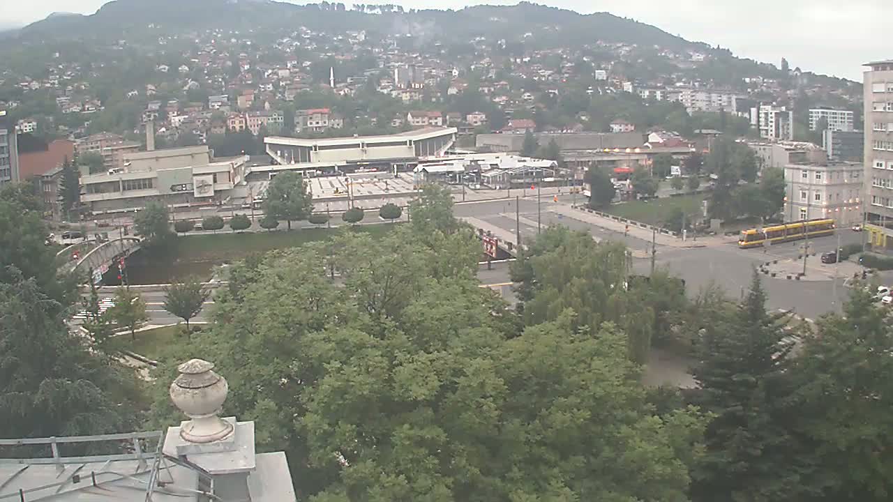 A high-angle view shows a city square with a bus station, a tram, and surrounding buildings on an overcast day.