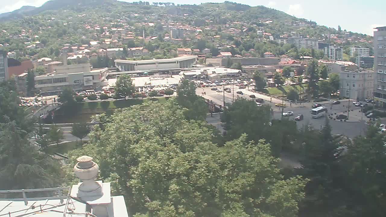 A high-angle view of a city on a sunny day, showing buildings, roads with vehicles, a large plaza, and a river flowing through a green space.