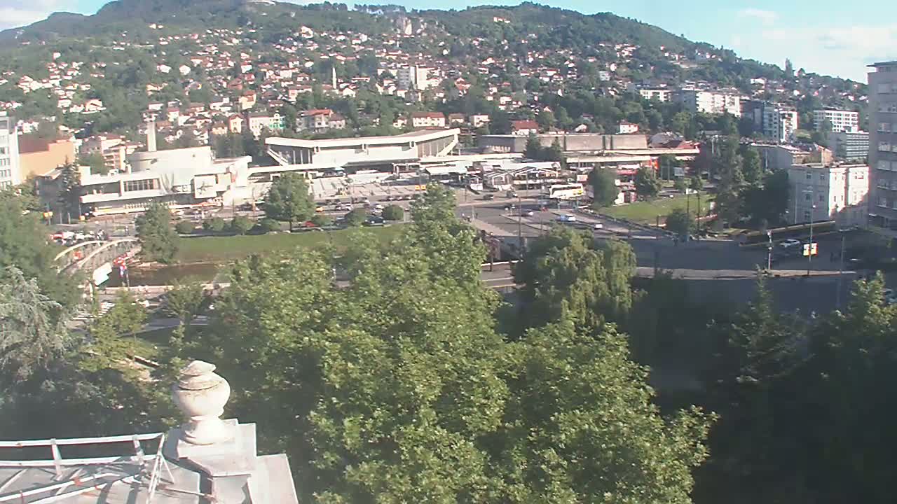 A high-angle, long shot on a sunny day shows a large city sprawled across a hillside, with a bus station and surrounding roads visible in the foreground, and green trees partially obscuring the view.