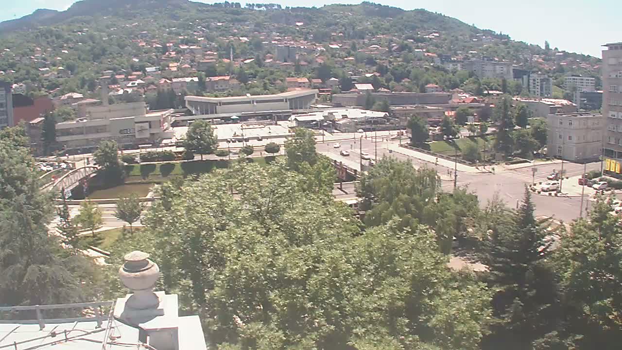 A high-angle view on a sunny day shows a city nestled against a hillside, with a river, bridge, and buildings including a large, flat-roofed structure, surrounded by green trees.